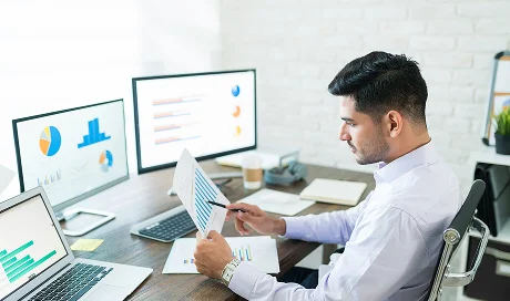 This image is used for DzLink - A man reviewing a printed document with a pen while sitting at a desk with two monitors and a laptop displaying business charts and data