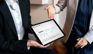 Image for DzIntel: Two people in business attire look at a tablet displaying a line graph with a downward trend.
