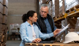 Two warehouse managers, a woman and an older man, reviewing documents and a laptop - DzTrack Centralized Inventory Asset Management
