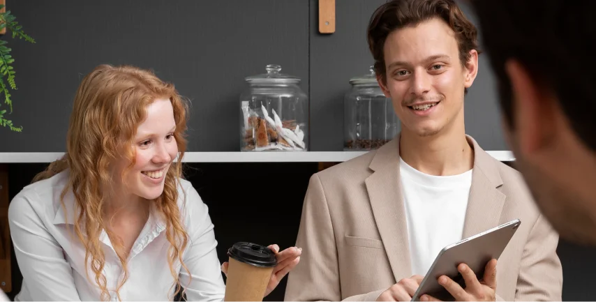 Two smiling coworkers, a woman with a cup of coffee and a man holding a tablet, sitting at a desk