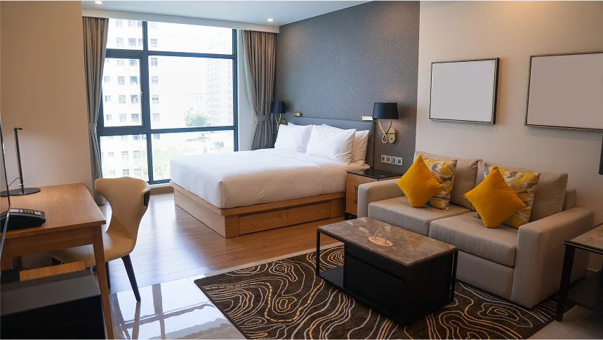 Modern hotel suite featuring a large bed, a sofa with yellow pillows, a marble coffee table, and a floor-to-ceiling window